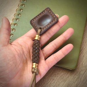 May include: A brown leather keychain with a braided leather handle and gold-coloured accents. The rectangular tag is stitched with a lighter brown thread. The keychain is attached to a tan cord.
