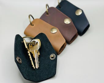 May include: A collection of leather keychains in various colors: black, brown, tan, and dark brown. Each keychain features a metal snap closure and a metal ring for attaching keys. One keychain is open, displaying several keys.