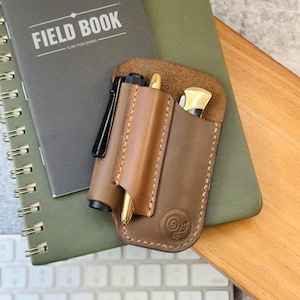 May include: A brown leather pen and knife holder with a gold pen and a knife with a gold handle. The holder is next to a gray notebook with the words "FIELD BOOK" and a green notebook. The holder has a decorative swirl design.
