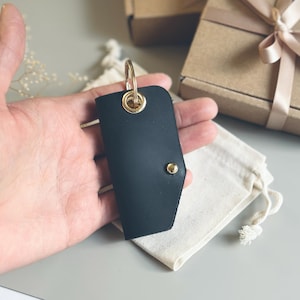 May include: Black leather key fob with a gold-colored metal ring and rivet. The key fob is held in a hand, with a small drawstring bag and wrapped gift boxes in the background. The key fob is a simple, elegant design.
