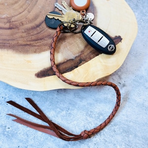 Puede incluir: Un llavero trenzado de cuero marrón con un mando de coche y varias llaves metálicas. El llavero tiene un lazo en el extremo con tres borlas de cuero. Las llaves y el llavero están sobre una superficie de madera.