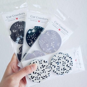 May include: A collection of car coasters in various colors, each featuring a leaf pattern. The coasters are packaged in clear plastic and labeled "Car Coasters Handmade with love."