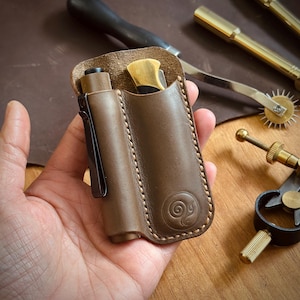 May include: A brown leather knife and flashlight sheath with a decorative embossed design. The sheath has a pocket for a small knife and a flashlight with a black clip. The sheath is stitched with a light brown thread.