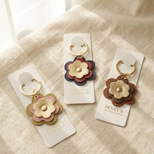 May include: Three leather flower keyrings in gold, pink, and brown. Each keyring has a gold-coloured ring and a small tag with the text "PENNY'S LEATHER HOUSE". The keyrings are displayed on a neutral-coloured surface.