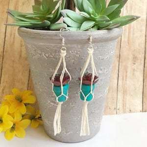 May include: A pair of handmade earrings featuring small, teal-colored pots with miniature succulent plants. The pots are suspended by cream-colored macrame, with a tassel at the bottom. The earrings are displayed in front of a gray pot with a succulent plant.