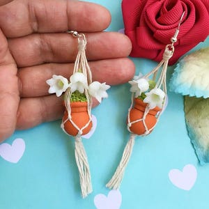 May include: A pair of handmade earrings featuring miniature orange terracotta pots with white flowers and green moss. The pots are suspended in macrame hangers with silver-colored ear wires. The background is a light blue with white heart shapes.