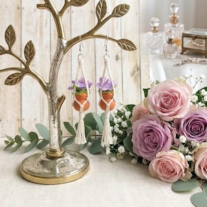 May include: A pair of handmade earrings featuring small terracotta pots filled with purple flowers, suspended in macrame holders. The earrings hang from a decorative gold tree-shaped jewelry stand. A bouquet of pink and purple roses is in the background.