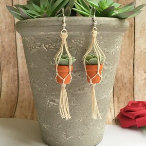 May include: A pair of dangle earrings featuring miniature potted succulents hanging from macrame plant hangers. The earrings are made with natural materials and have a rustic, bohemian style.