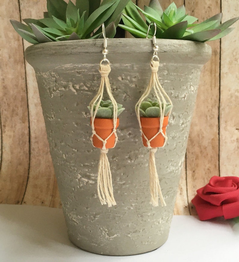 May include: A pair of dangle earrings featuring miniature potted succulents hanging from macrame plant hangers. The earrings are made with natural materials and have a rustic, bohemian style.