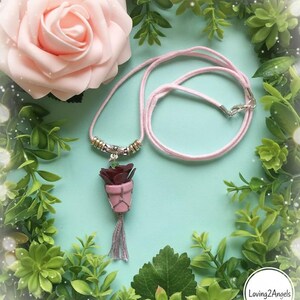 May include: A pink cord necklace with a pendant featuring a burgundy rose in a pink pot with a gray tassel. The necklace has silver-colored metal accents. A large pink rose and greenery surround the necklace.
