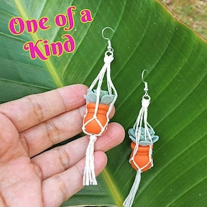 May include: A pair of dangle earrings featuring miniature orange terracotta-style pots with green succulent plants held in white macrame hangers with tassels. The text One of a Kind is printed in yellow with a pink outline in the upper left corner.