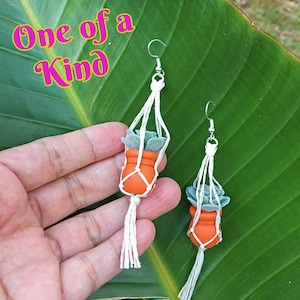 May include: A pair of handmade earrings featuring miniature terracotta pots with green succulent plants hanging from macrame string. The earrings are a one-of-a-kind design.