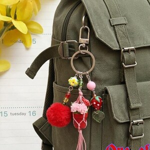 May include: An olive green canvas backpack featuring a silver metal keychain with multiple charms, including a red pom-pom, a miniature potted plant in a macrame hanger, and a red strawberry bead. The text One of a kind is visible in red.
