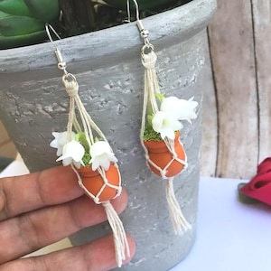May include: Handmade earrings featuring miniature terracotta pots with white flowers and green moss. The pots are suspended in macrame hangers with silver-tone ear wires, creating a unique, nature-inspired accessory.