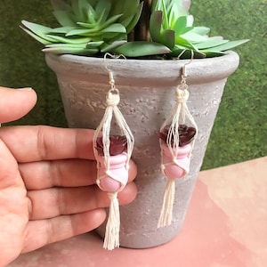 May include: A pair of handmade earrings featuring miniature pink and burgundy succulent plant pots suspended in macrame holders. The earrings have silver-colored hooks and white tassels, with a succulent plant in each pot.