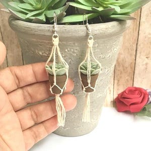 May include: A pair of dangle earrings featuring miniature brown pots with light green succulents, each held in a cream-colored macrame hanger with a tassel at the bottom, suspended from silver-toned metal earring hooks for pierced ears.