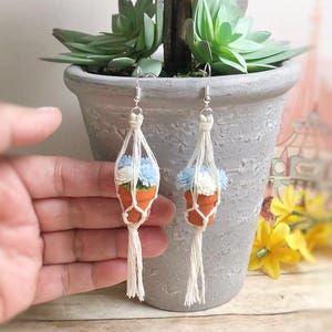 May include: A pair of dangle earrings featuring miniature terracotta-colored pots held in off-white macrame hangers. Each pot contains small, textured blue and white flowers. The earrings are attached to silver-toned metal hooks for hanging.