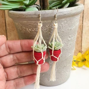 May include: A pair of dangle earrings featuring miniature red pots containing light green succulents, each suspended in a cream-colored macrame hanger made of knotted cord, finished with silver-toned metal earring hooks for pierced ears.