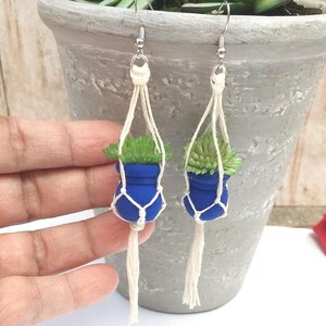 May include: A pair of handmade earrings featuring miniature blue planters with green succulent plants. The planters are suspended in macrame-style holders with dangling tassels. The earrings have silver-colored ear wires.