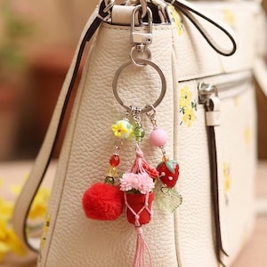May include: A cream-colored textured handbag with a yellow floral pattern features a multi-charm keychain. The keychain includes a red pom-pom, a miniature pink potted plant in a macrame hanger, and a red strawberry charm with green leaves.