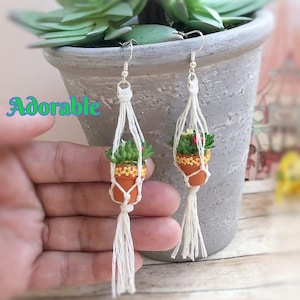 May include: A pair of dangle earrings featuring miniature terracotta-colored pots with floral patterns, each held in a white macrame hanger with a tassel. The pots contain small green succulents. The word Adorable is printed in blue and green.