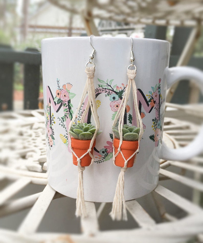May include: A pair of earrings featuring small potted succulents hanging from macrame plant hangers. The earrings are made with natural twine and have a tassel detail. The succulents are a light green color and are potted in small orange pots.