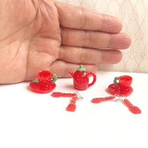 May include: A miniature red teapot, two matching teacups on saucers, and two sets of tiny red forks and spoons. Each piece features a white dotted strawberry pattern and small green accents. The items are arranged on a white surface.