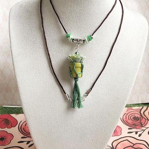 May include: A handcrafted necklace featuring a pendant shaped like a small succulent plant in a yellow pot, wrapped with green twine. The necklace has a brown cord and a green tassel. The necklace is displayed on a white mannequin.