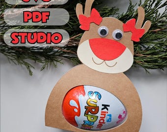 May include: A reindeer-shaped gift box made of brown cardstock, holding a Kinder Surprise egg. The reindeer has googly eyes, a red nose, and red bow accents. The text "SVG PDF STUDIO" is in the upper left corner.