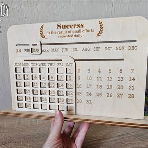 K&ouml;nnte beinhalten: Ein h&ouml;lzerner Desktop-Kalender mit einem monatlichen Kalenderraster und einem Zitat, das lautet "Erfolg ist das Ergebnis kleiner Anstrengungen, die t&auml;glich wiederholt werden". Der Kalender ist lasergeschnitten und bereit zum Zusammenbauen.
