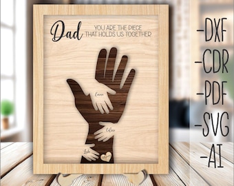 May include: A laser-cut wooden sign with the text "Dad You Are The Piece That Holds Us Together". The sign features a large hand with smaller hands inside, each labeled with a name: Luke, Emma, and Celine. The sign is on a wooden surface.