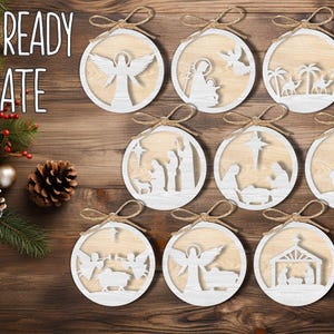 May include: Laser-cut wooden Christmas ornaments with various nativity scenes, including angels, the holy family, and the manger. Each ornament is round with a light wood center and a white border, tied with a twine bow. The words "LASER READY TEMPLATE" are in the upper left.