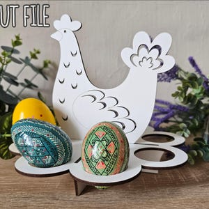 May include: White wooden chicken egg holder with six egg slots. The chicken is holding a green and red decorated egg in its beak. The holder is on a wooden surface with green plants in the background. The text "LASER CUT FILE" is in the top left corner of the image. The text "DXF - CDR - PDF - SVG - AI" is in the top right corner of the image.