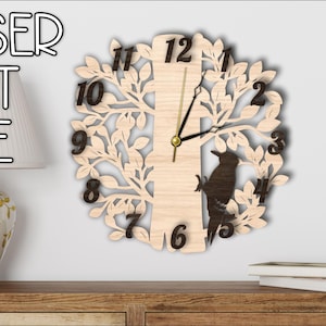 Puede incluir: Reloj de pared de madera cortado con láser con diseño de árbol y pájaro. El reloj tiene números y manecillas negras. El texto "LASER CUT FILE" es visible. El reloj se muestra en una pared.