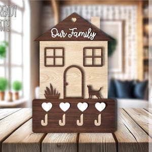 May include: A laser-cut wooden house-shaped key holder with the text "Our Family" on the front. The house has four heart-shaped hooks and a small dog silhouette below the door. The key holder is on a wooden table.