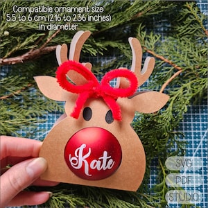 Op de afbeelding: Een bruin rendierornament met een rode strik en een rode bal met de naam "Kate" in witte letters. Het ornament is ontworpen om een bal te bevatten met een diameter van 5,5 tot 6 cm. De afbeelding bevat ook de tekst "SVG", "PDF" en "STUDIO".