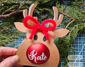 May include: A brown reindeer ornament with a red bow and a red ball with the name "Kate" in white script. The ornament is designed to hold a ball that is 5.5 to 6 cm (2.16 to 2.36 inches) in diameter. The image also includes the text "SVG", "PDF", and "STUDIO".