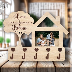 May include: Wooden key holder with a house design and a tree with the text "Home is not a place it's a feeling". The house has a photo frame with a family of four inside. There are five key hooks and a heart shape above each hook.