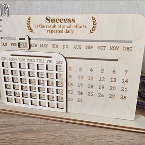 K&ouml;nnte beinhalten: Ein h&ouml;lzerner Schreibtischkalender mit einem monatlichen Kalenderrraster und einem motivierenden Zitat: "Erfolg ist das Ergebnis kleiner Anstrengungen, die t&auml;glich wiederholt werden."