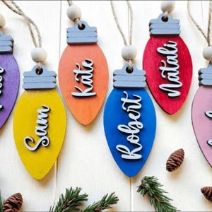 May include: Colorful wooden Christmas ornament cutouts in the shape of light bulbs, each with a name in white script. The ornaments are purple, yellow, orange, blue, red, and pink. Each ornament has a gray top and a wooden bead for hanging.