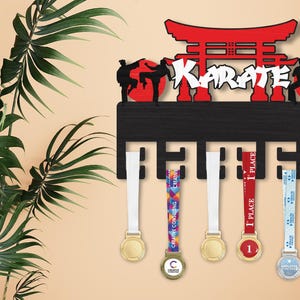 Puede incluir: Expositor de medallas de karate de madera negra con detalles rojos. Presenta la palabra "Karate" y siluetas de karatecas. Incluye medallas con cintas coloridas, ideal para mostrar logros.