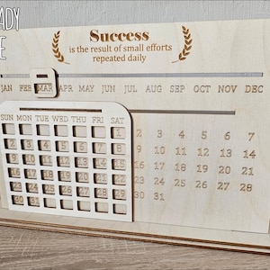 K&ouml;nnte beinhalten: Eine lasergeschnittene Holzkalender-Vorlage mit einem Zitat &uuml;ber Erfolg. Der Kalender verf&uuml;gt &uuml;ber ein monatliches Raster mit Wochentagen und den Zahlen 1-31. Das Zitat lautet "Erfolg ist das Ergebnis kleiner Anstrengungen, die t&auml;glich wiederholt werden".