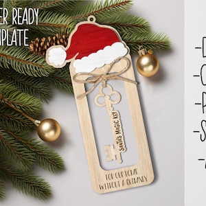 Könnte beinhalten: Ein hölzerner Weihnachtsschmuck mit einem Weihnachtsmützen-Design. Der Schmuck zeigt einen Schlüssel mit der Aufschrift "Santa's Magic Key" und den Text "For our home without a chimney". Umgeben von Tannenzweigen, goldenen Kugeln und einem Tannenzapfen.