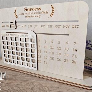 K&ouml;nnte beinhalten: Ein lasergeschnittener Holz-Desktop-Kalender mit einem monatlichen Kalenderrraster und einem Zitat, das lautet "Erfolg ist das Ergebnis kleiner Anstrengungen, die t&auml;glich wiederholt werden". Der Kalender ist so konzipiert, dass er zusammengebaut und personalisiert werden kann.