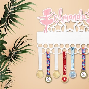 May include: A white medal holder with the name "Amanda" and ballerina figures. The holder displays several medals with colorful ribbons. Green leaves are in the background.