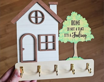 May include: Wooden key holder with a house and tree design. The house is white with brown trim and a brown roof. The tree is green with a watercolor effect. The key holder has five hooks. The text on the tree reads "Home is not a place it's a feeling."