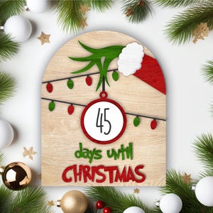 May include: A wooden Christmas countdown sign featuring a green Grinch hand holding a red and white Santa hat. The sign has a white circle with the number 45 and the text "days until Christmas".