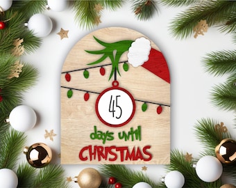 May include: A wooden Christmas countdown sign with a green Grinch hand holding a red and white Santa hat. The sign has a white circle with the number 45 and the text "days until Christmas".