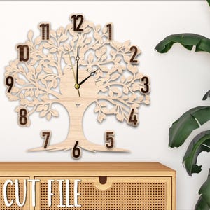 May include: A laser cut wooden clock in the shape of a tree with intricate branches and leaves. The clock face is in the centre of the tree with Roman numeral hour markers. The clock is on a wooden shelf with a wicker drawer.