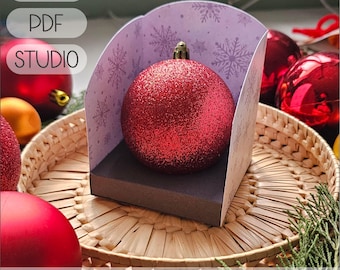 May include: A red glitter Christmas ornament inside a decorative box with a snowflake pattern. The box is designed to hold ornaments 2.55 to 2.75 inches in diameter. The image also shows the text "SVG", "PDF", and "STUDIO".